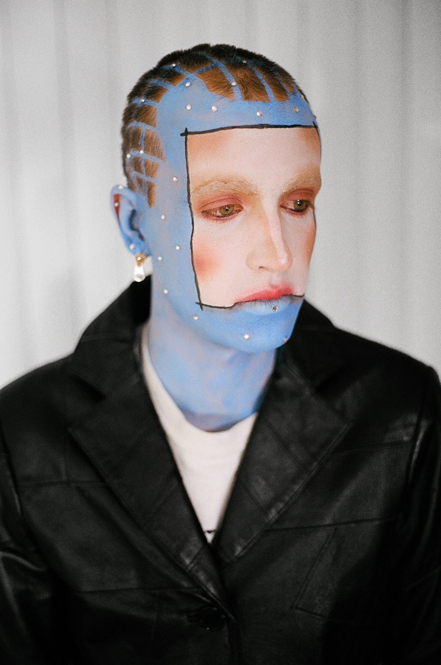 Close up photo of a model looking downwards longingly with blue skin and and a white square in the center of their face wearing a black leather jacket in front of a white background